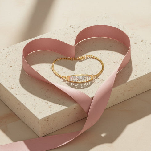 A 14k gold-filled bracelet and a pink ribbon shaped into a heart rest on a speckled stone slab in soft light.