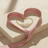A 14k gold-filled bracelet and a pink ribbon shaped into a heart rest on a speckled stone slab in soft light.