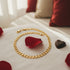 A gold necklace, rose petals, and a 14k gold-filled anklet rest on a white bed with red and beige pillows in the background.