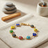 A 14k gold-filled evil eye bracelet with colorful charms rests on beige fabric, beside stones and burning incense.