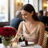 A woman in an elegant dress smiles at a table with roses, champagne, and a 14k gold filled Mariner link necklace in a jewelry box.