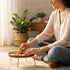 A woman sits cross-legged, arranging crystals and an Evil Eye charm on a wooden table in a cozy, sunlit room filled with incense.