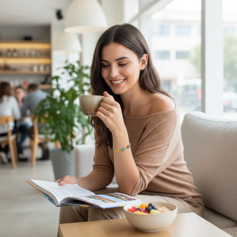 A woman sits on a couch, smiling while drinking coffee, wearing her 14k Gold-Filled Evil Eye Bracelet, and reading a magazine.