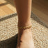 A close-up of a 14k gold-filled Cuban curb link anklet on an ankle, sunlight illuminating the woven rug and wooden floor.