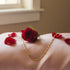 A red rose, petals, and a 14k gold filled mariner link necklace rest on soft pink fabric near a sunlit window.