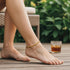 A person wearing a 14k gold-filled anklet sits outdoors on a wooden deck with an iced drink nearby, surrounded by lush greenery.