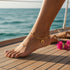 A woman's foot with a 14k Gold-filled Anklet rests on a boat deck near the sea, with pink flowers and sandals in the background.