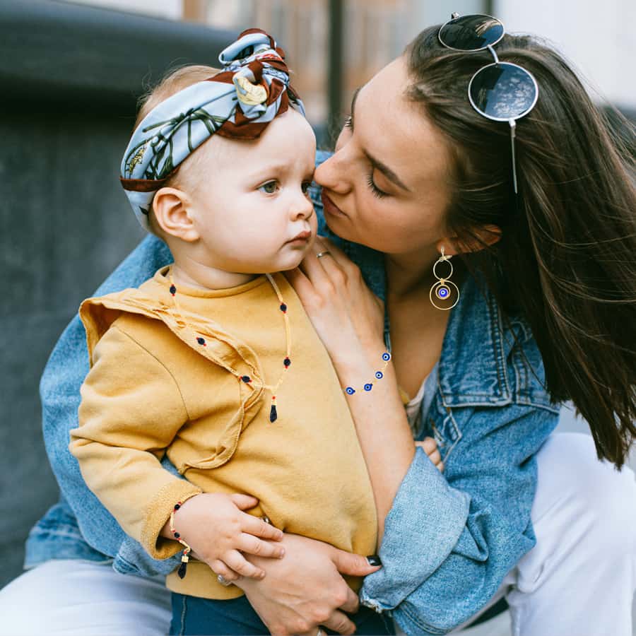 A woman is holding a baby while wearing an Azabache Bracelet and Necklace, some blue evil eye Jewelry, sunglasses and a denim jacket