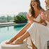 Two women in white dresses sit by a pool, laughing and toasting pink drinks, each wearing a stylish 14k gold-filled anklet.