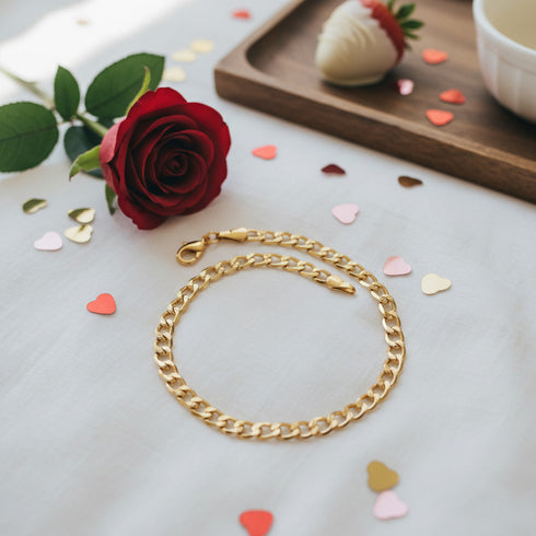 Gold chain bracelet on a white surface with heart confetti, a red rose, and a 14k gold-filled Cuban curb link anklet nearby.