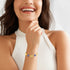 A woman smiles in a white sleeveless top, wearing the Blue Evil Eye Charm Bracelet and Heart Lucky Chain Rolo Link on her wrist—a stylish good luck piece she shows off while gently touching her chin.