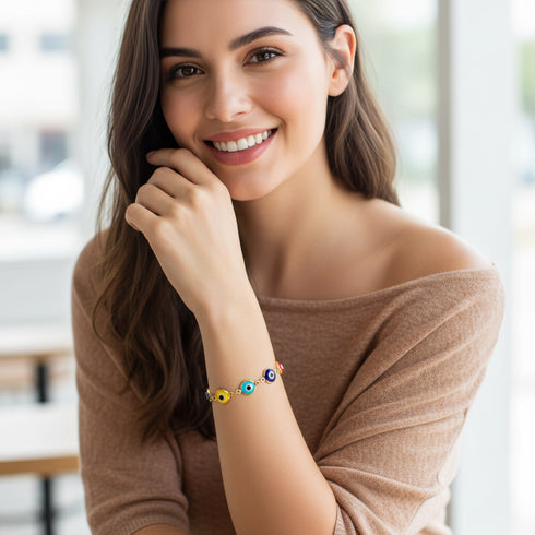 A smiling woman with long brown hair wears a beige off-shoulder top and an Evil Eye Bracelet, sitting indoors.