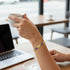 A hand wearing a 14k Gold-Filled Bracelet holds a white cup near a laptop on a wooden café table.