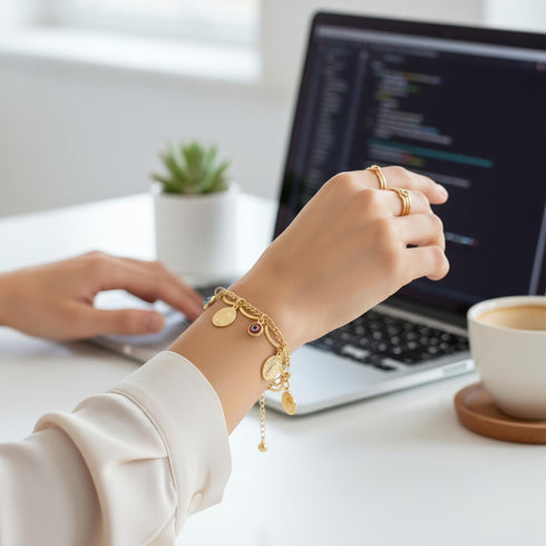 A person wearing a 14k Gold-filled bracelet types on a laptop at a desk with a plant, coffee cup, and code visible on the screen.