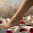 A gold Figaro Chain Anklet with charms graces her bare foot, resting on white satin and rose petals against a floral background.