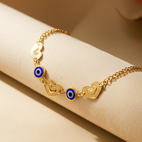 The Blue Evil Eye charm bracelet and Heart Lucky chain rolo link features heart charms and blue evil eye beads on a 14k gold-filled chain, offering a shiny finish for style, protection, and good luck. Displayed on light beige fabric.
