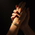 Two hands gently touching, one adorned with a sparkling 14k gold-filled bracelet against a dark background.