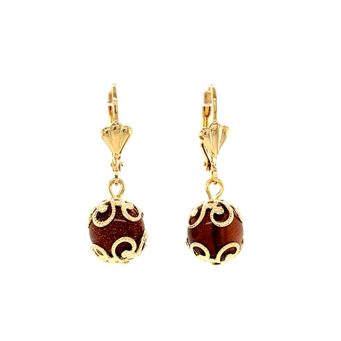 The Adorned Goldstone Beads Drop Earrings are crafted with 14k gold-filled materials and feature exquisite brown goldstone beads nestled in ornate gold filigree designs. These earrings come with leverback closures and offer a smooth, polished finish for an elegant appearance, all while being lead and nickel free to accommodate sensitive ears.