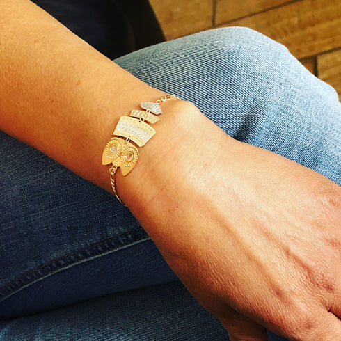 A person wearing the WISE OWL 2-Tones Gold Filled Charm Bracelet, featuring a blend of gold and silver nature-inspired charms, gently rests their hand on denim jeans, highlighting this elegant piece of jewelry.
