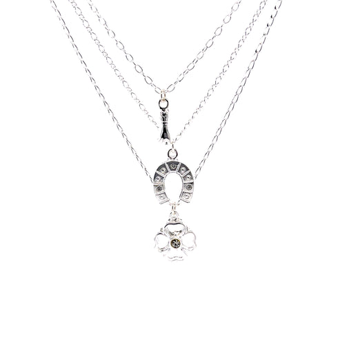 The Good Luck Charms Pendant Stacked Necklace, measuring 17 inches and silver plated, features a beautiful layered design with charms including a horseshoe, a small Eiffel Tower, and a floral motif accented by a central stone. These enchanting symbols hang elegantly on delicate silver chains against a pristine white backdrop.