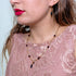 A woman in a pink lace top showcases Azabache jewelry, including an exquisite gold necklace and matching earrings enriched with red and black gemstones. These 14k gold filled pieces are renowned for their spiritual protection qualities, as highlighted by the Genuine Jet Black Azabache Bracelet and Necklace for Evil Eye, making her ensemble both elegant and meaningful.