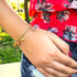 A hand adorned with a Mega Lucky Charm Bracelet 11 Amulets Good Luck and Evil Eye Protection rests on a knee. The person, exuding positive energy, is wearing a floral top and denim shorts. In the background, grass and a tree complete the serene scene.