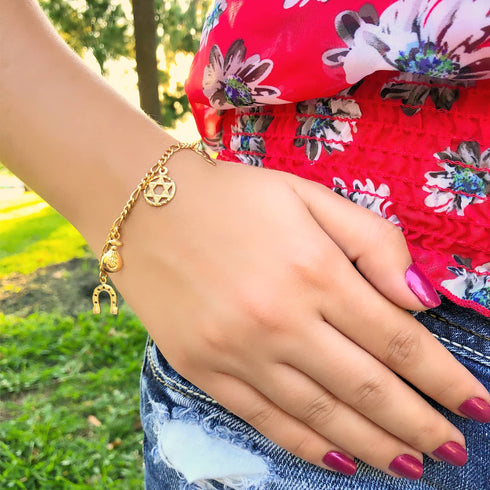 A hand with pink nail polish rests on denim jeans, showcasing a Women Mega Lucky Charm Bracelet—an exquisite 14k Gold-filled accessory designed with 7 amulets for good luck and evil eye protection, including a star and horseshoe. The individual wears a floral red top, surrounded by greenery and sunlight.