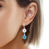 The image showcases a person wearing the Blue Evil Eyes Silver Plated and Zirconia Stones Dangle Earrings, featuring a circular silver design embellished with zirconia stones and accented by a dangling blue evil eye motif. The person's short brown hair and ear are highlighted against the simple white background.
