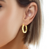 A close-up shot captures a single 14k gold-filled hoop earring with a textured design from the product "14k Gold Filled Hoops Earrings 1 Inch Round Textured Design," glimmering elegantly as it hangs from the ear. Light brown hair frames the accessory against a plain white backdrop.