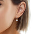 Close-up of a person's ear wearing Beautiful Drop Zirconia Stone Huggie earrings in rose gold. These earrings include a gold hoop embellished with small diamonds and a larger square-cut zirconia stone pendant, designed to be safe for all skin types. The individual’s short hair is tucked behind the ear.