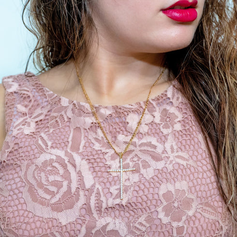 Wearing a pink lace dress, a woman elegantly displays the Large Iced Crucifix Pendant Necklace, featuring Figaro links and adorned with zirconia stones. Her bright red lips and loosely styled hair enhance the soft, light-hued background.