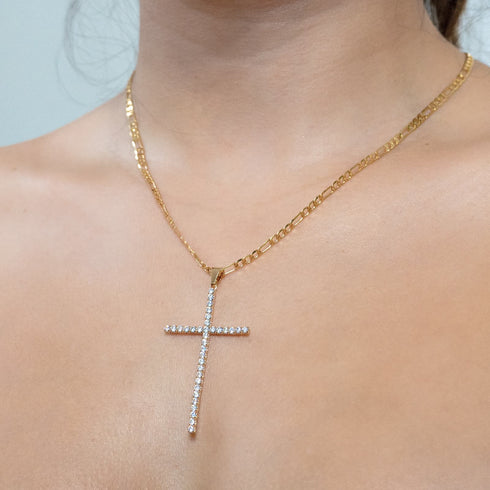 A person wearing the Large Iced Crucifix Pendant Necklace with Figaro Link, featuring a 14k gold-filled chain adorned with sparkling zirconia stones. The neutral background highlights the elegance of this jewelry piece.